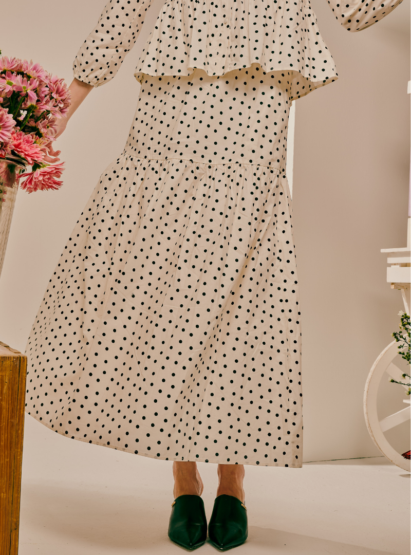 A woman wearing a long cream maxi dress with a black polka dot pattern, featuring a ruffled peplum-style bodice and a tiered skirt. She is wearing dark green pointed-toe mules.