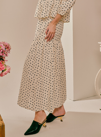 A model wearing a cream-colored maxi dress with a black polka dot pattern, paired with dark green pointed-toe mule heels with gold accents, in a minimalist studio setting.