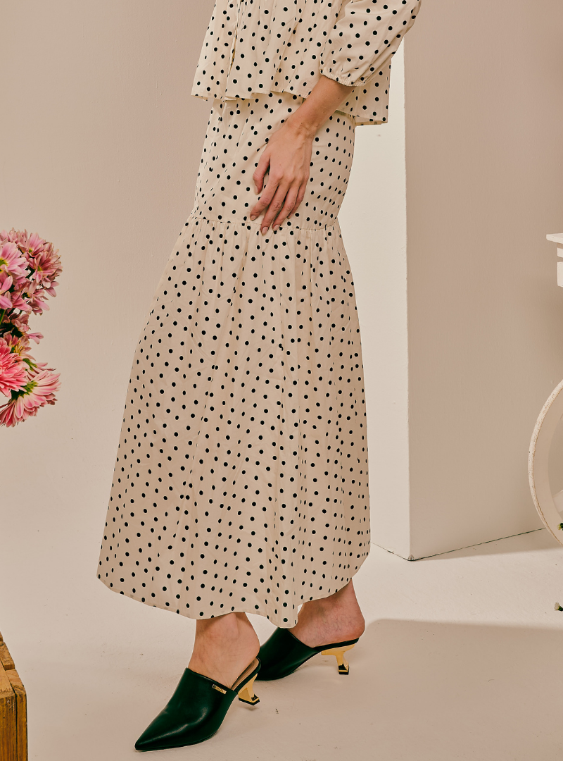 A model wearing a cream-colored maxi dress with a black polka dot pattern, paired with dark green pointed-toe mule heels with gold accents, in a minimalist studio setting.