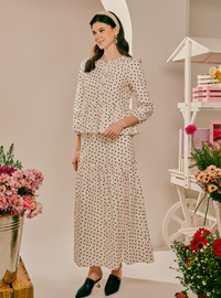 A woman models a matching two-piece dress set featuring a cream base with a black polka dot pattern. The set includes a long-sleeve top with a peplum waist and a matching long skirt.