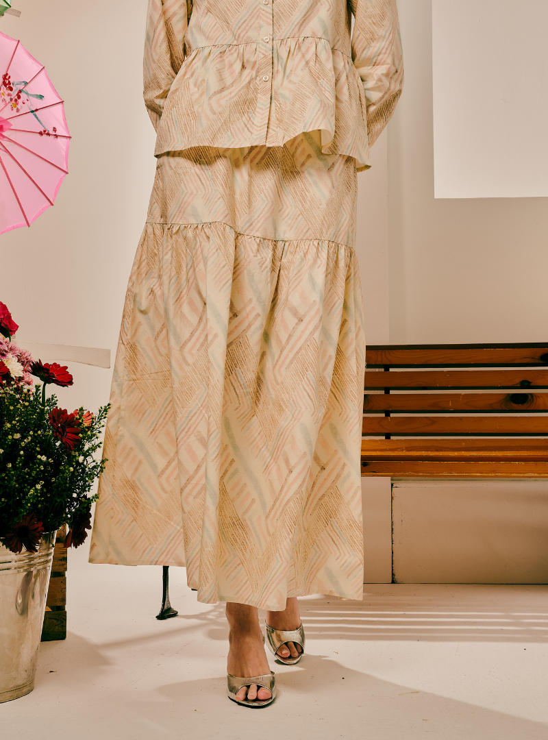 A model wearing a beige and gold patterned two-piece set consisting of a long-sleeve peplum top and a matching tiered maxi skirt, paired with metallic silver sandals. The setting is indoors with a wooden bench and a pink parasol visible in the background.
