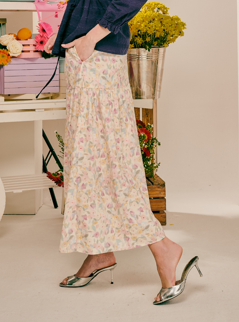 A person wearing a long cream maxi skirt with a delicate pastel floral print, styled with a navy blue top and metallic silver mule heels, standing in a studio setting with decorative flowers in the background.