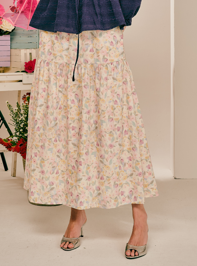 A model wearing a tiered A-line midi skirt with a delicate pastel floral print on a cream background, paired with a navy blue ruffled top and metallic silver heeled sandals.