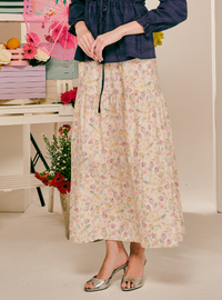 A model wearing a long, tiered maxi skirt with a delicate pastel floral print on a cream background, paired with a navy blue top and metallic silver heeled sandals.