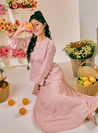A woman wearing a pink long-sleeve maxi dress with small white polka dots sits on a white floor surrounded by flowers, baskets of lemons and oranges, and a mirror.