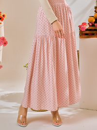 A model wearing a pink tiered maxi skirt with a small black polka dot pattern, styled with a beige long-sleeve top and clear heeled sandals.