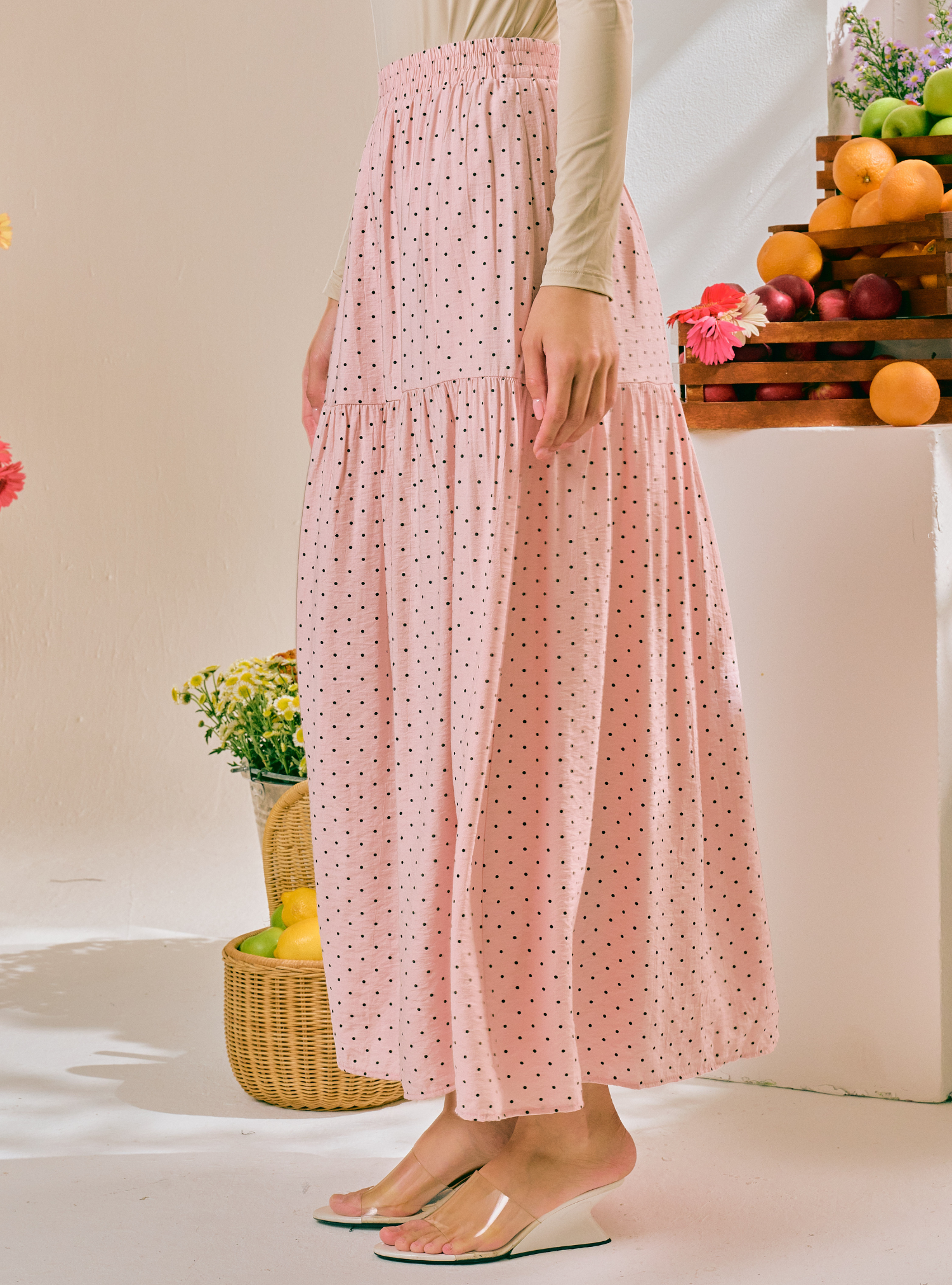A model wearing a pink tiered maxi skirt with a small black polka dot pattern, paired with a beige long-sleeve top and clear slide sandals, set against a warm, sunlit studio backdrop with decorative fruit and flower props.