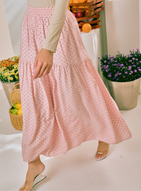 A woman wearing a light pink tiered maxi skirt with a small black polka dot pattern, styled with a beige long-sleeve top and clear heeled sandals in a bright, styled indoor setting.