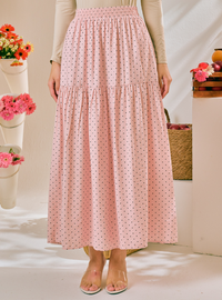 A pink tiered maxi skirt with a small black polka dot pattern, worn by a model in a styled indoor setting with flowers and baskets.