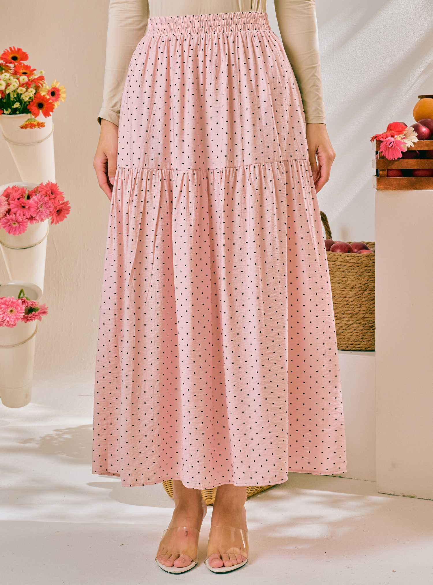 A pink tiered maxi skirt with a small black polka dot pattern, worn by a model in a styled indoor setting with flowers and baskets.