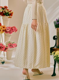 A woman wearing a cream-colored, tiered maxi skirt with a small black polka dot pattern, styled with a beige long-sleeve top and white wedge sandals. The background is a warm, softly lit studio setting decorated with colorful flower arrangements.