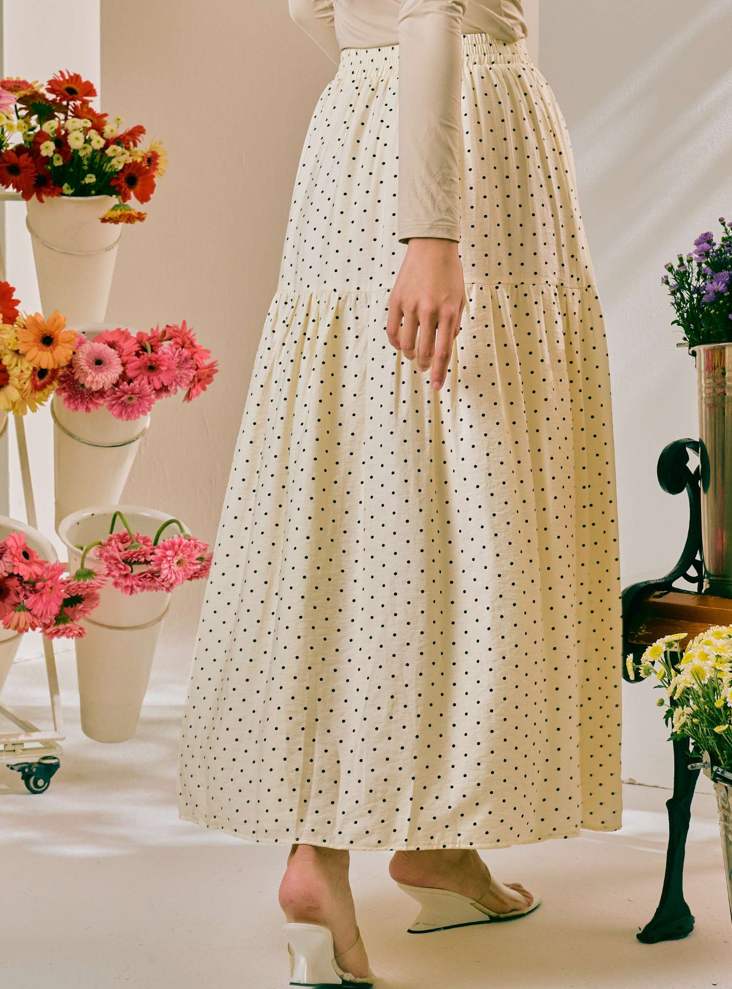 A woman wearing a cream-colored, tiered maxi skirt with a small black polka dot pattern, styled with a beige long-sleeve top and white wedge sandals. The background is a warm, softly lit studio setting decorated with colorful flower arrangements.