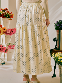 A cream-colored, tiered maxi skirt with a small black polka dot pattern, worn by a model standing in a brightly lit room decorated with potted flowers.