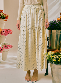 A woman wearing a cream-colored, tiered maxi skirt with a small black polka dot pattern, paired with a beige long-sleeve top and beige flat sandals. The background is a bright, sunlit studio decorated with potted flowers.