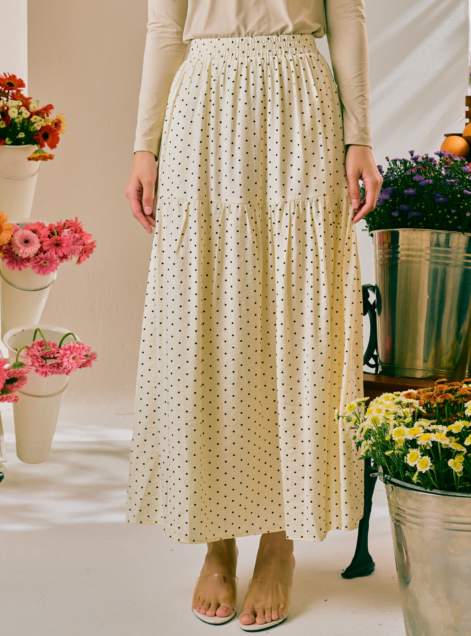 A woman wearing a cream-colored, tiered maxi skirt with a small black polka dot pattern, paired with a beige long-sleeve top and beige flat sandals. The background is a bright, sunlit studio decorated with potted flowers.
