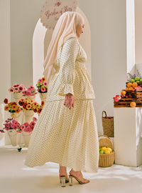 A woman wearing a cream-colored, long-sleeved maxi dress with a small white floral print and a matching hijab, standing in a bright, styled indoor setting with flowers and fruit.