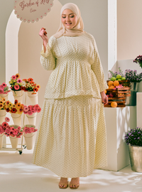 A woman wearing a cream-colored two-piece modest dress with a small green polka dot pattern and a matching hijab, standing in a brightly lit indoor garden setting with potted flowers and fruit.