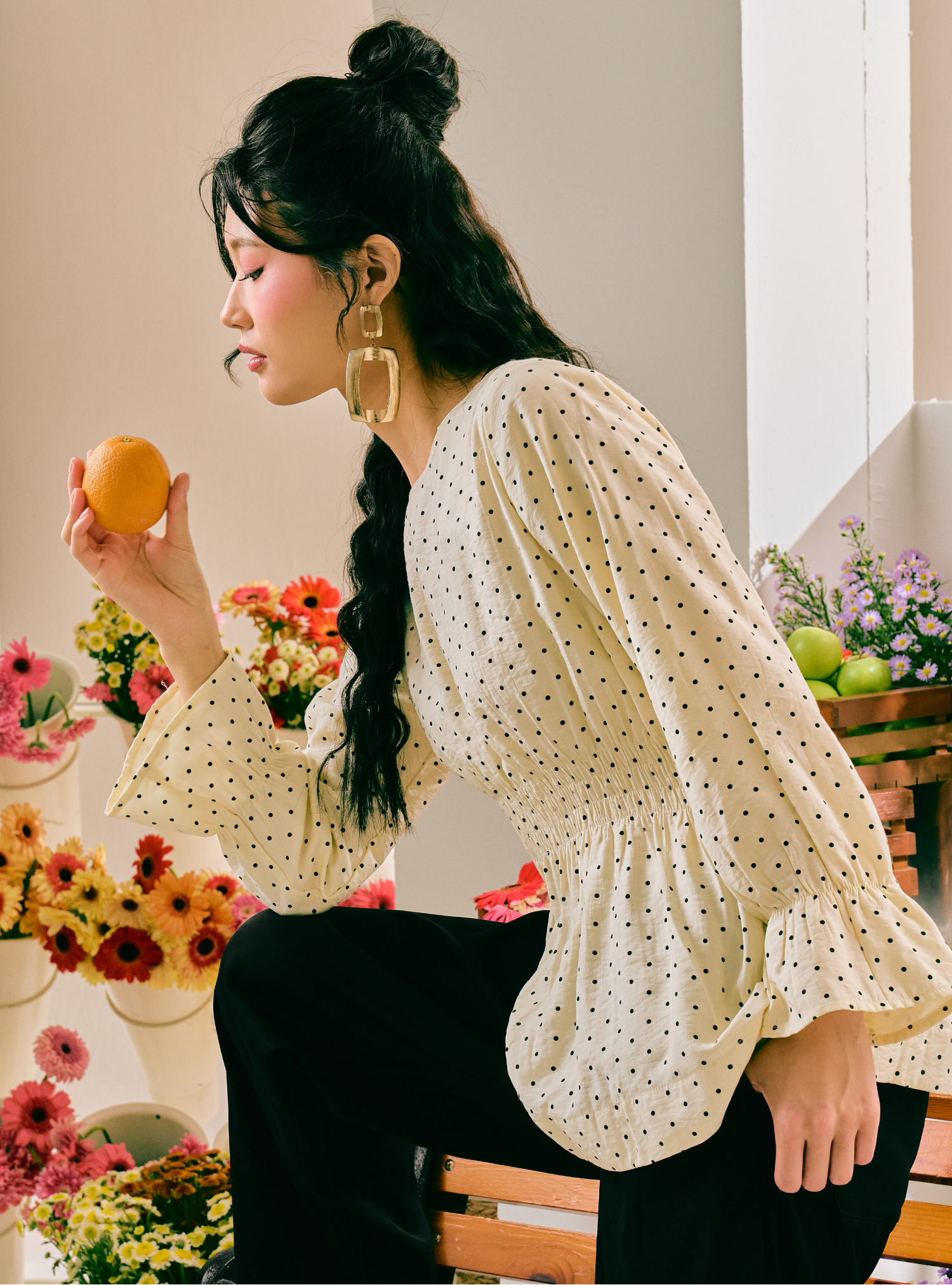 A woman with dark hair in a high bun and side braid, wearing a cream-colored long-sleeve blouse with a small black polka dot pattern and black pants, holding an orange while looking to the side. She is seated on a wooden bench in a brightly lit room decorated with flowers.
