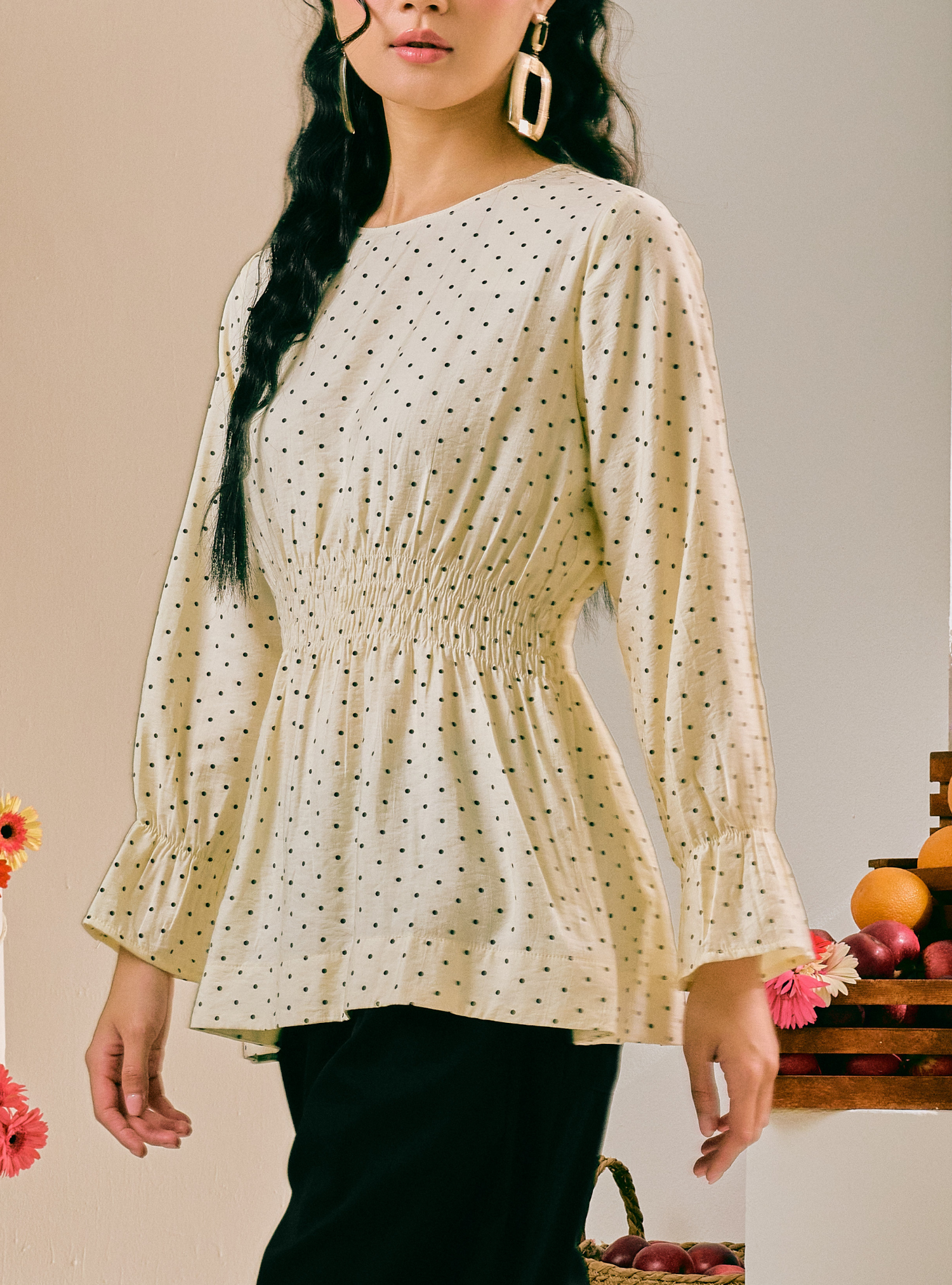 A woman wearing a cream-colored long-sleeve blouse with a black polka dot pattern and a smocked waist, paired with black pants.