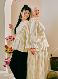 Two women modeling matching cream-colored polka dot outfits in an indoor setting with a warm beige background. One woman wears a long-sleeve peplum top with black pants, and the other wears a matching long-sleeve peplum top with a beige hijab and a long skirt.