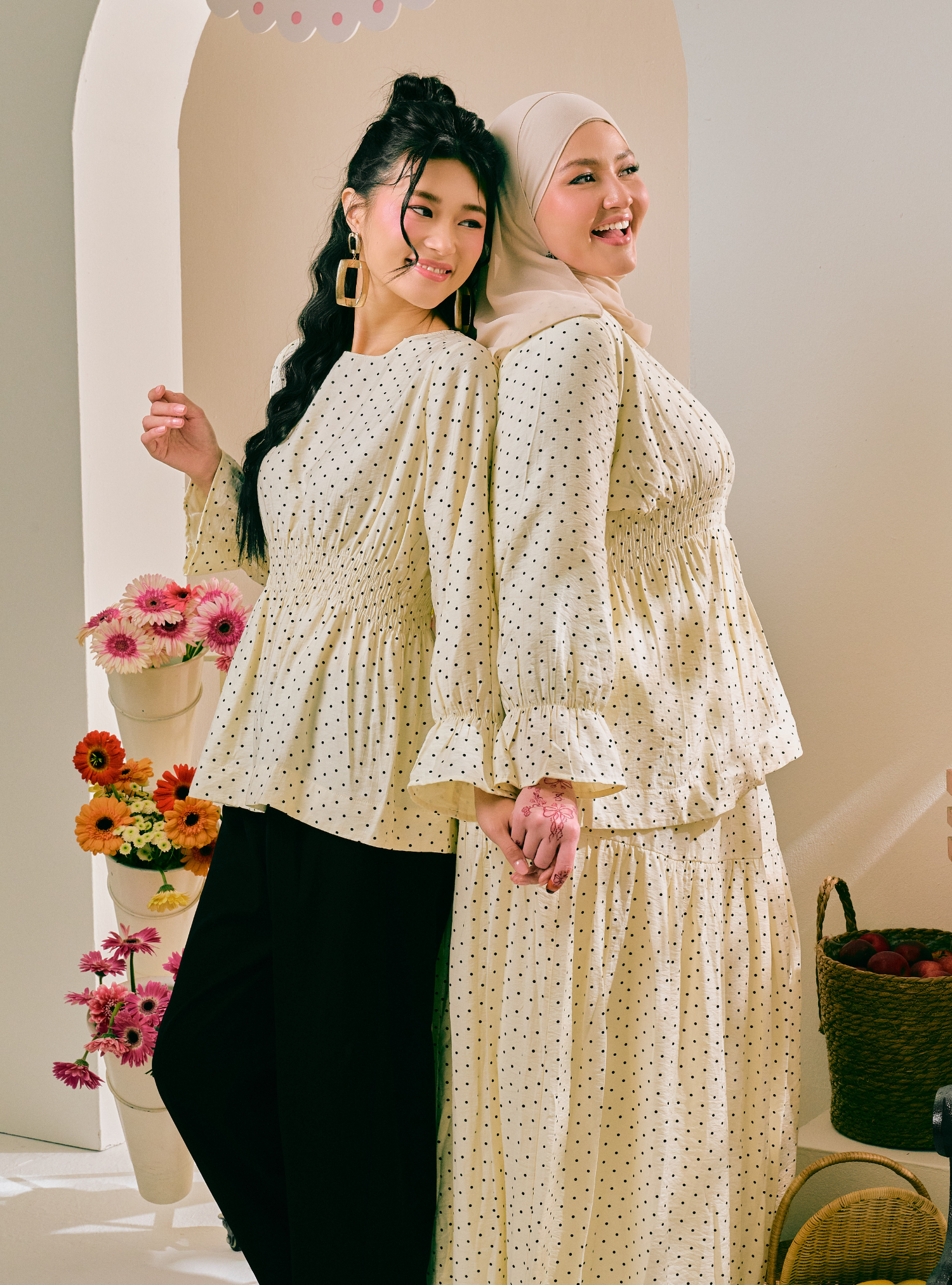 Two women modeling matching cream-colored polka dot outfits in an indoor setting with a warm beige background. One woman wears a long-sleeve peplum top with black pants, and the other wears a matching long-sleeve peplum top with a beige hijab and a long skirt.