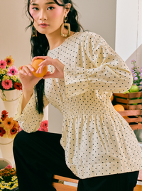 A woman wearing a cream-colored long-sleeve blouse with a black polka dot pattern and black pants, holding an orange in a styled indoor setting with flowers and fruit.