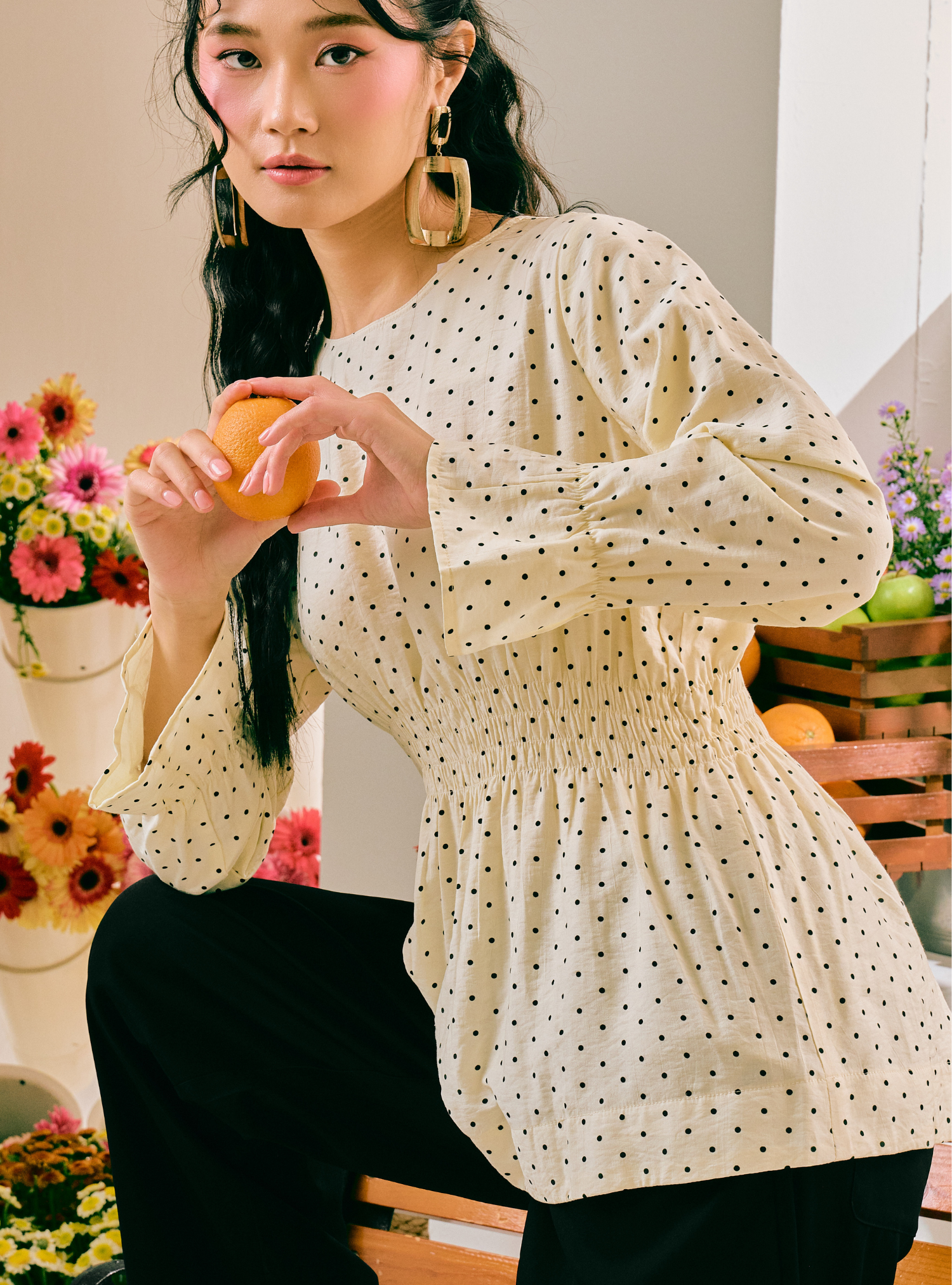 A woman wearing a cream-colored long-sleeve blouse with a black polka dot pattern and black pants, holding an orange in a styled indoor setting with flowers and fruit.