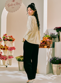 A woman stands in a bright boutique interior wearing a cream-colored, long-sleeve, textured crochet top with a small white pattern and black wide-leg pants. The background features white display stands with flowers, fruit, and a sign that reads 'Garden of Her'.