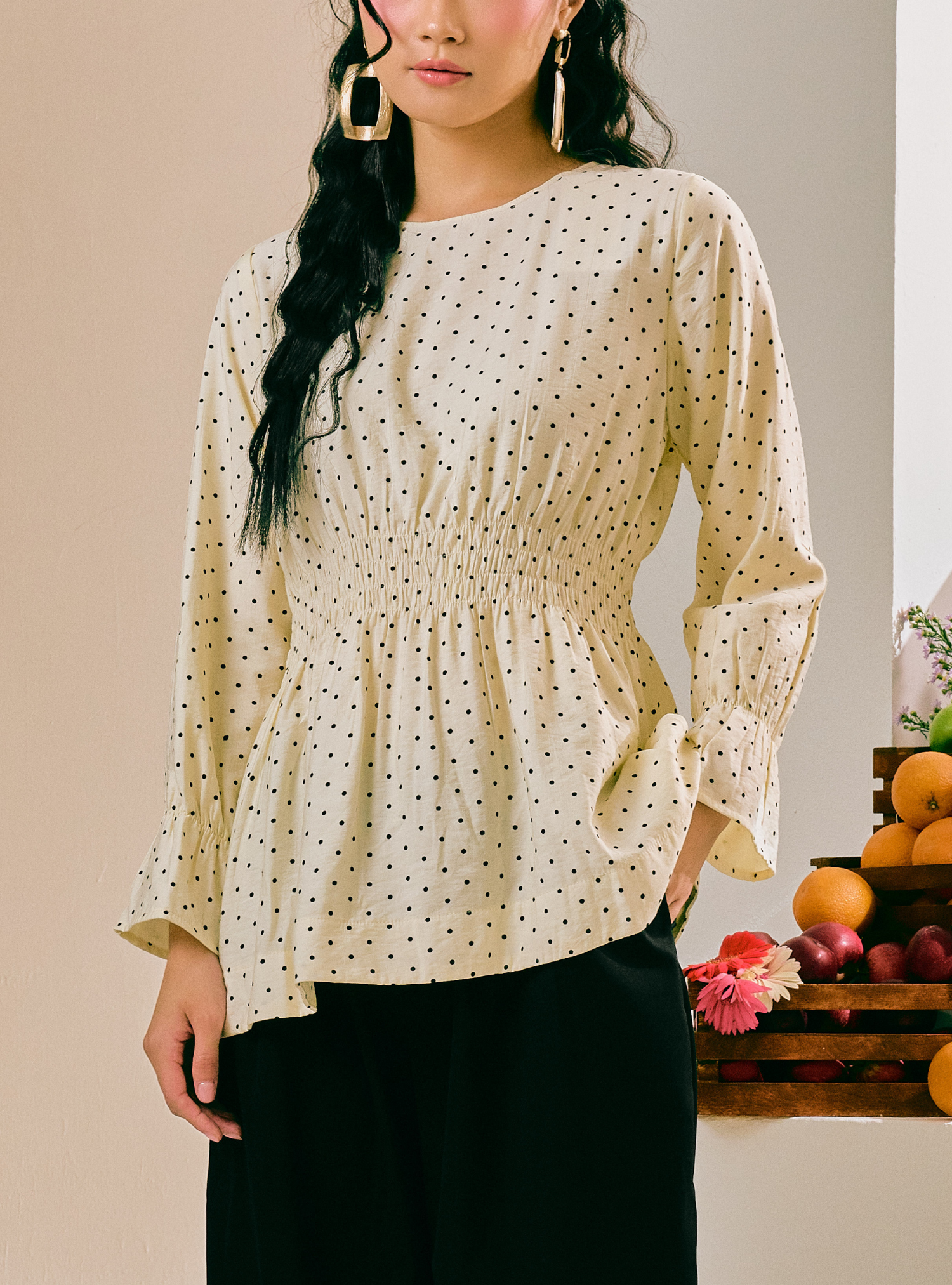 A woman wearing a cream-colored long-sleeve blouse with a small black polka dot pattern and a smocked waist, paired with black pants.