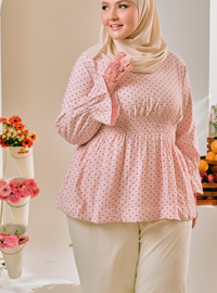 A woman wearing a light pink long-sleeve blouse with a small black polka dot pattern and a beige hijab, standing in an indoor setting with decorative flowers in the background.