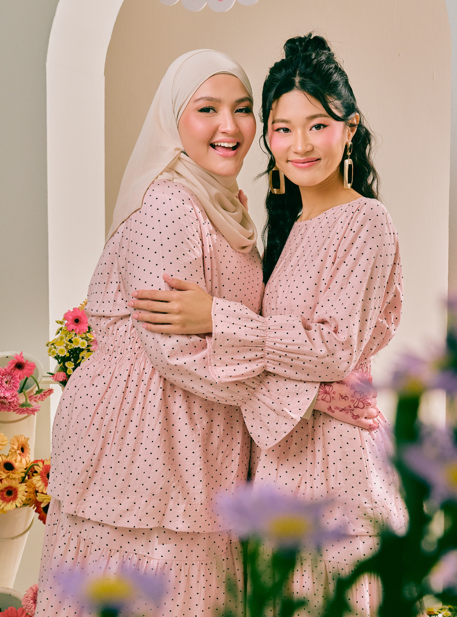 Two women embracing and smiling, both wearing matching light pink long-sleeve dresses with a small dark polka dot pattern. One woman wears a light beige hijab.