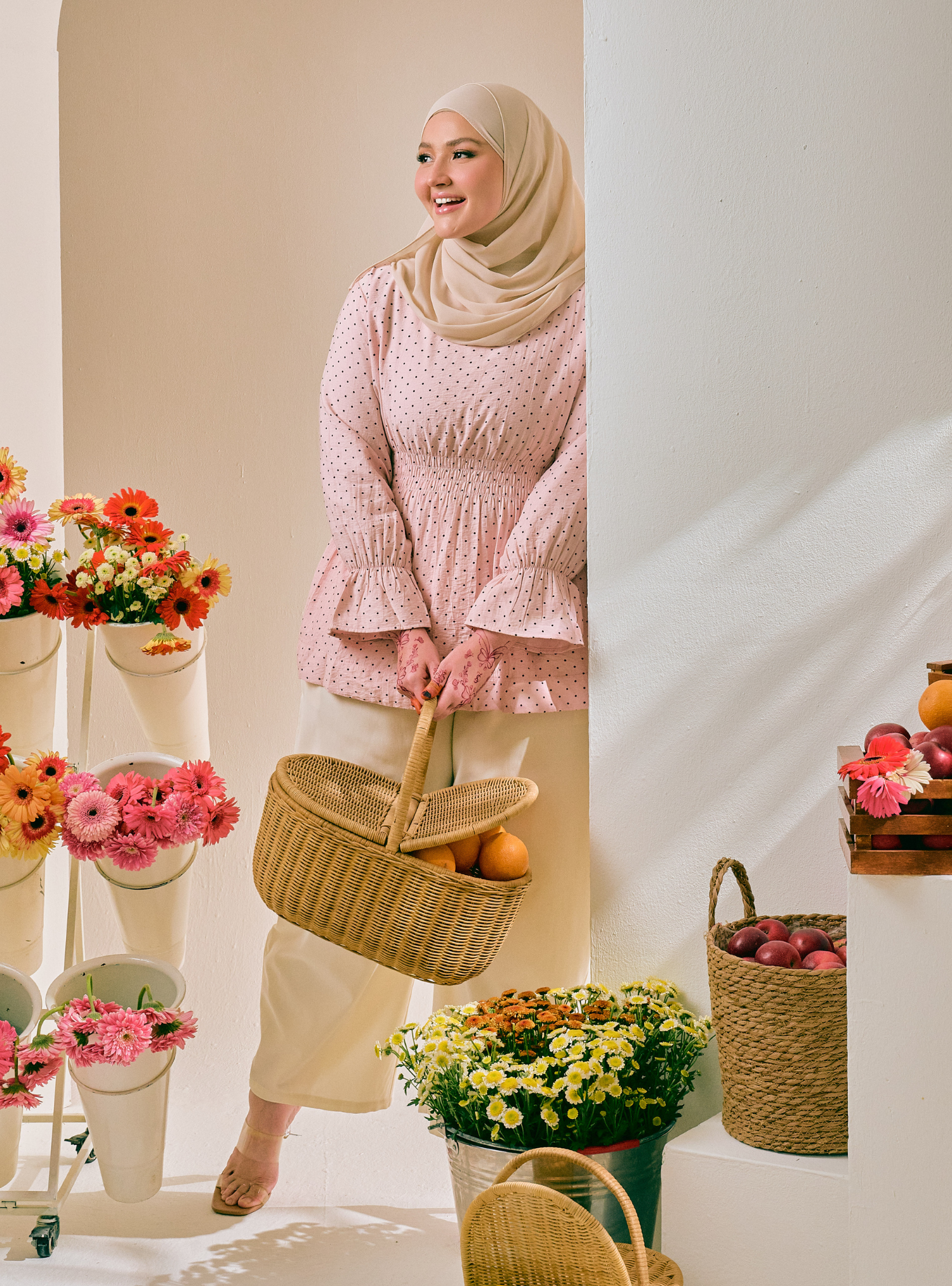 A woman wearing a beige hijab and a pink patterned long-sleeve blouse with cream pants stands in a bright studio setting, holding a woven basket. The scene includes floral arrangements and additional baskets on white display platforms.