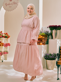 A woman wearing a light pink long-sleeve modest maxi dress with a small white polka dot pattern and a matching headscarf, standing in a bright, styled studio setting with flowers and fruit props.
