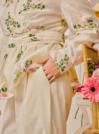 A close-up of a cream-colored kimono-style robe with pink and green floral embroidery, worn by a model whose hand is in a pocket. A wicker basket with pink flowers is visible on the right side.