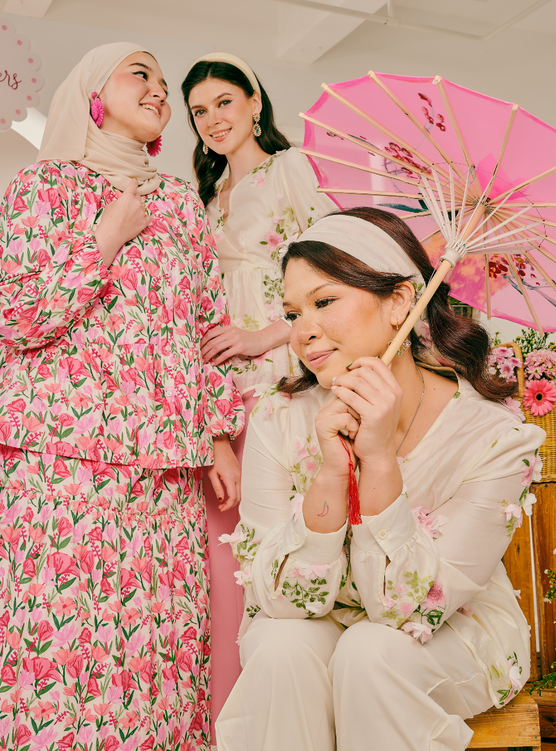 Three women posing together in coordinated floral traditional outfits. One woman on the left wears a pink and beige floral dress with a matching hijab, one seated woman in the foreground wears an ivory floral long-sleeve top and pants with a white headband and holds a pink parasol, and another woman stands behind them in a matching cream floral outfit.
