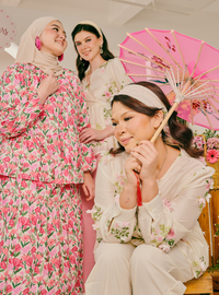 Three women posing together in coordinated floral traditional outfits. One woman on the left wears a pink and beige floral dress with a matching hijab, one seated woman in the foreground wears an ivory floral long-sleeve top and pants with a white headband and holds a pink parasol, and another woman stands behind them in a matching cream floral outfit.