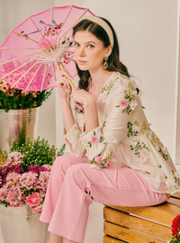 A woman sits on a wooden crate wearing a light pink and white floral print blouse with matching pink pants, holding a pink floral paper umbrella in a studio setting with flowers.