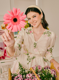 A woman wearing a cream-colored floral dress and a beige headband holds up a large pink gerbera daisy flower. She is seated in a basket filled with pink and white flowers, with a softly lit indoor background.