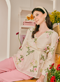 A woman wearing a cream-colored wrap-style blouse with pink and green floral embroidery, paired with pink pants, sitting in a decorated room with flowers and shelves.