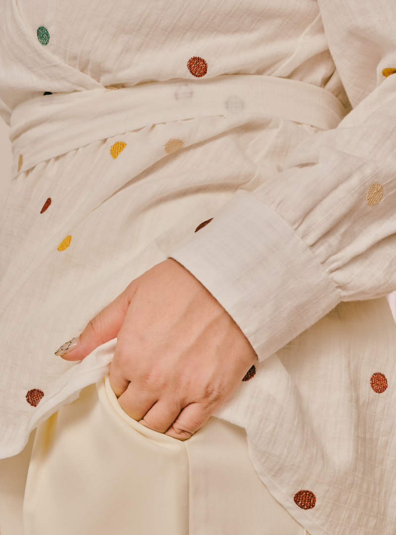 Close-up of a cream-colored cotton gauze kimono-style robe with colorful embroidered polka dots, featuring a matching tie belt and a hand reaching into a front pocket.