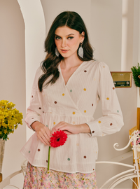 A woman wearing a cream-colored wrap blouse with multicolored polka dots, holding a red flower, set against a warm, softly lit indoor background.