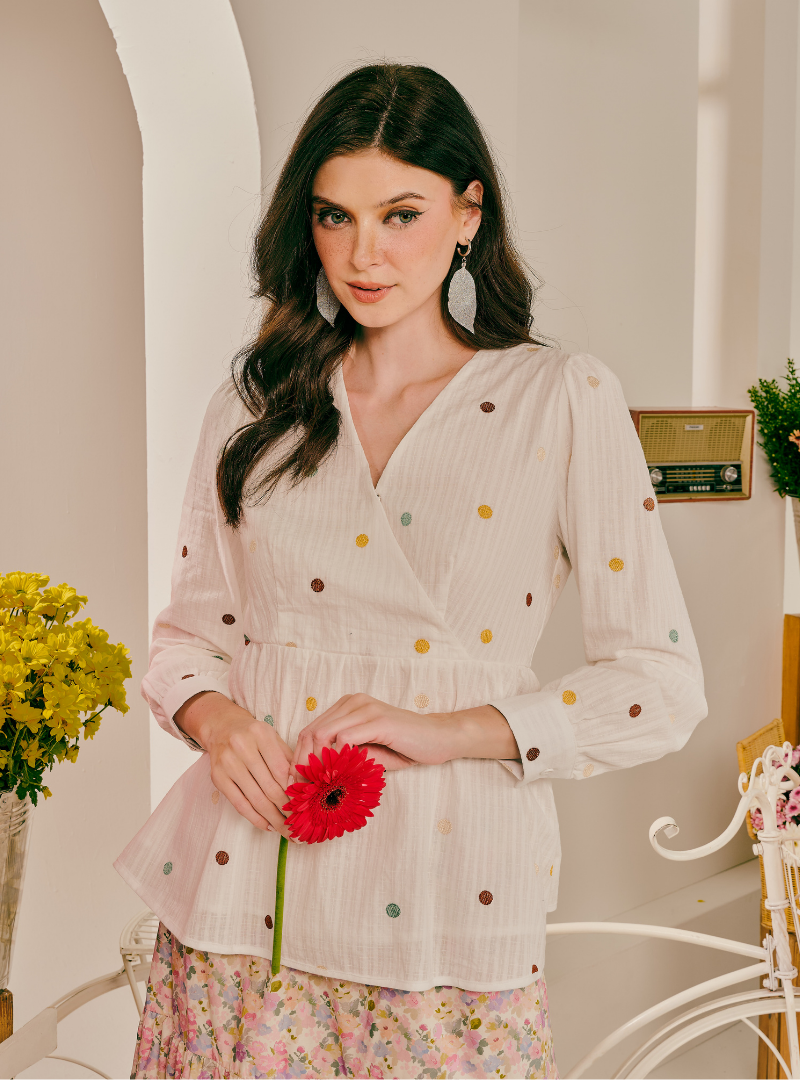 A woman wearing a cream-colored wrap blouse with multicolored polka dots, holding a red flower, set against a warm, softly lit indoor background.