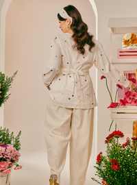 A woman is shown from the back wearing a cream-colored two-piece outfit with small colorful embroidered dots, featuring a long-sleeve top with a tie waist and matching wide-leg pants. She is standing in a styled studio setting with arches, shelves, and pink and red flowers.