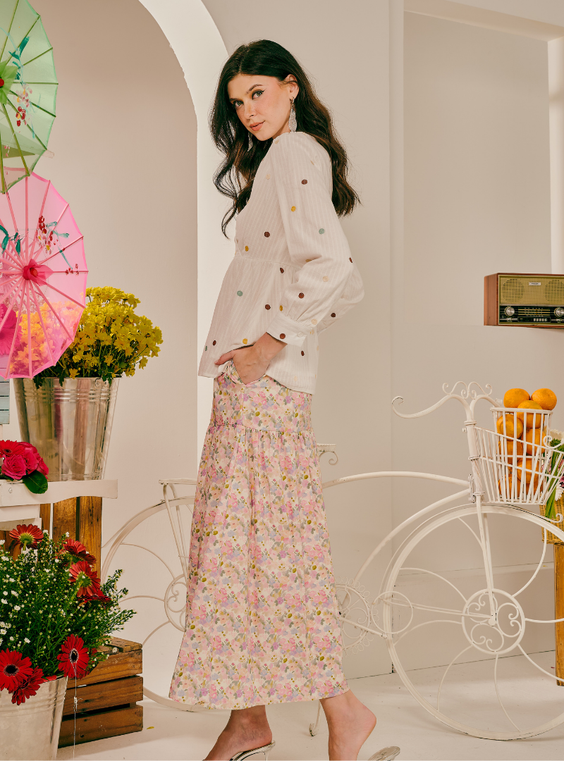 A woman stands in a bright indoor set wearing a white long-sleeve blouse with small colorful embroidered dots and a long pink floral skirt. Decorative umbrellas, flowers, and a white bicycle prop are visible in the background.