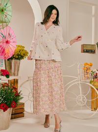 A woman models a two-piece outfit consisting of a white long-sleeve blouse with colorful embroidered dots and a matching pink floral midi skirt, styled in a bright, vintage-inspired room with a white bicycle prop.