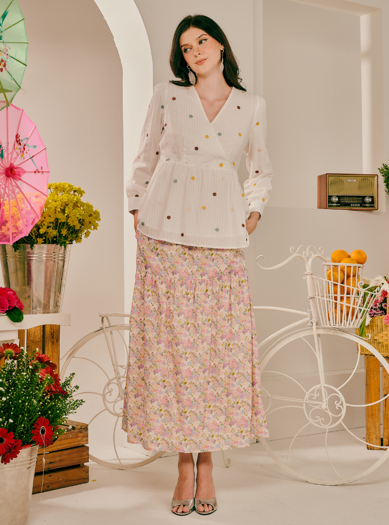 A woman stands in a styled indoor set wearing a white long-sleeve blouse with small multicolor embroidered dots and a long pink floral skirt. The scene includes decorative umbrellas, flowers, a vintage radio, and a white bicycle prop.