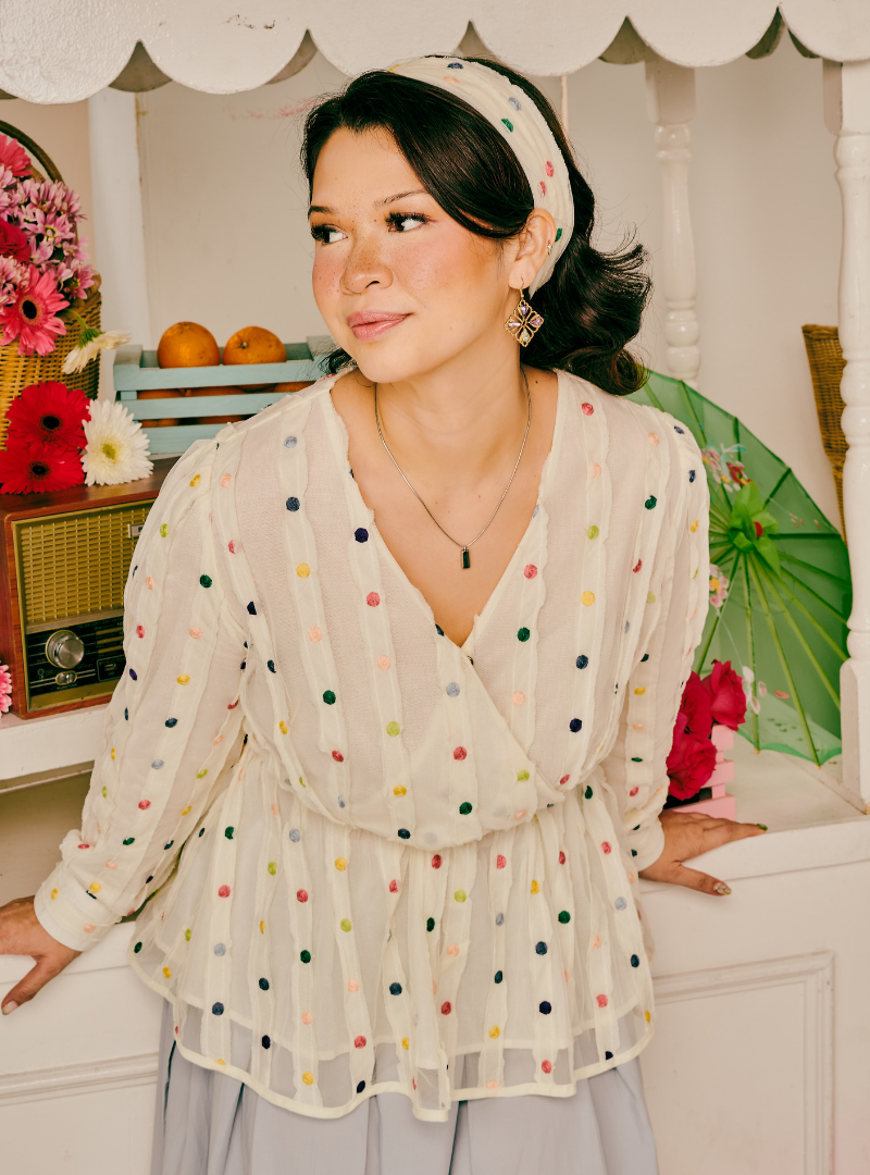 A woman wearing a cream-colored, long-sleeved wrap-style blouse with multicolored polka dots, paired with a matching polka-dot headband and a light gray skirt. She is posed in front of a white decorative screen with vintage props like a radio, flowers, and a green parasol.