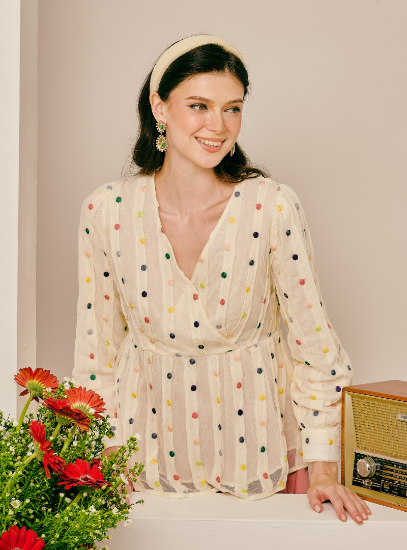 A woman wearing a cream-colored long-sleeve blouse with multicolored polka dots, styled with a cream headband and gold earrings, leaning against a wall next to a vintage radio and red flowers.