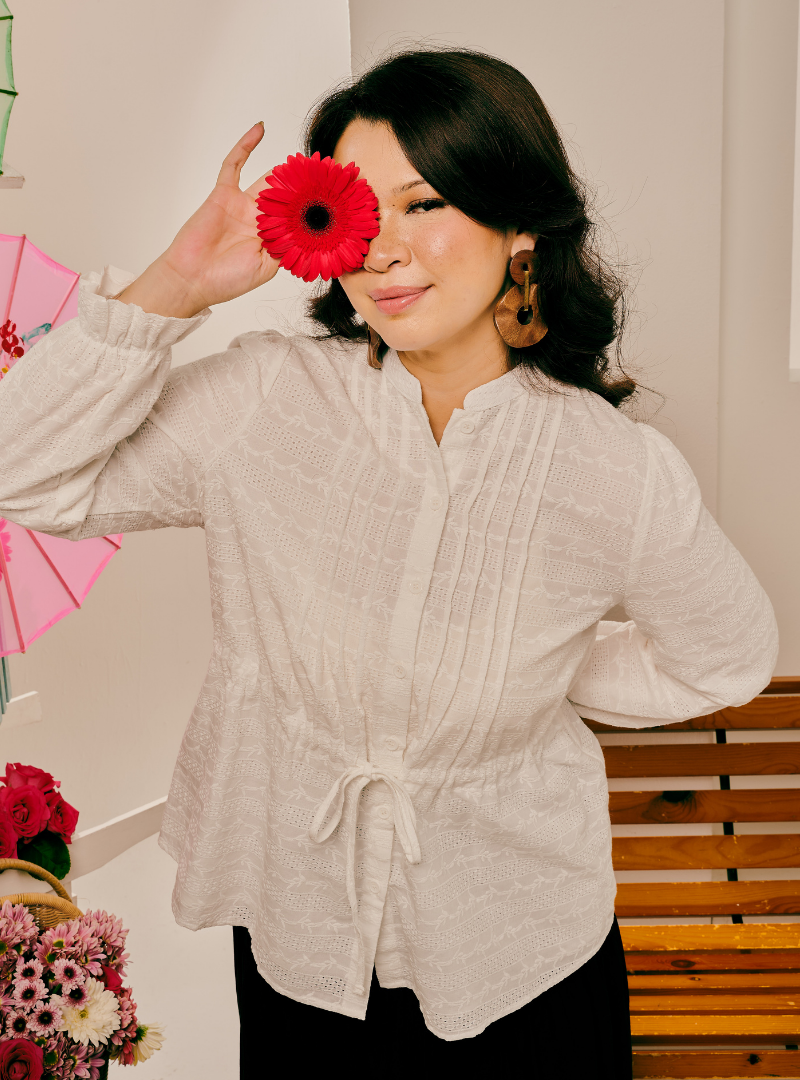 A woman wearing a cream-colored textured long-sleeve blouse with a drawstring waist, holding a red flower in front of one eye, set against an indoor background with a wooden bench.