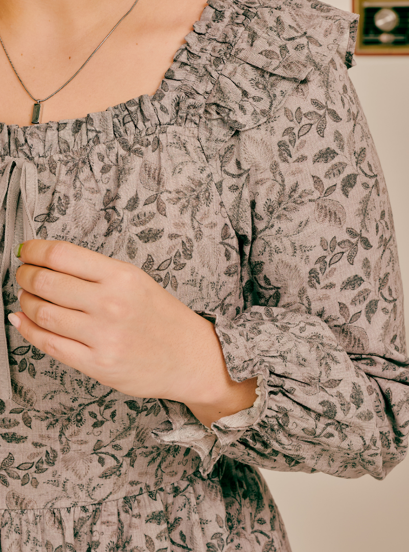 Close-up of a woman wearing a long-sleeve blouse with a gray and beige floral leaf pattern, ruffled neckline, and tie detail at the chest.